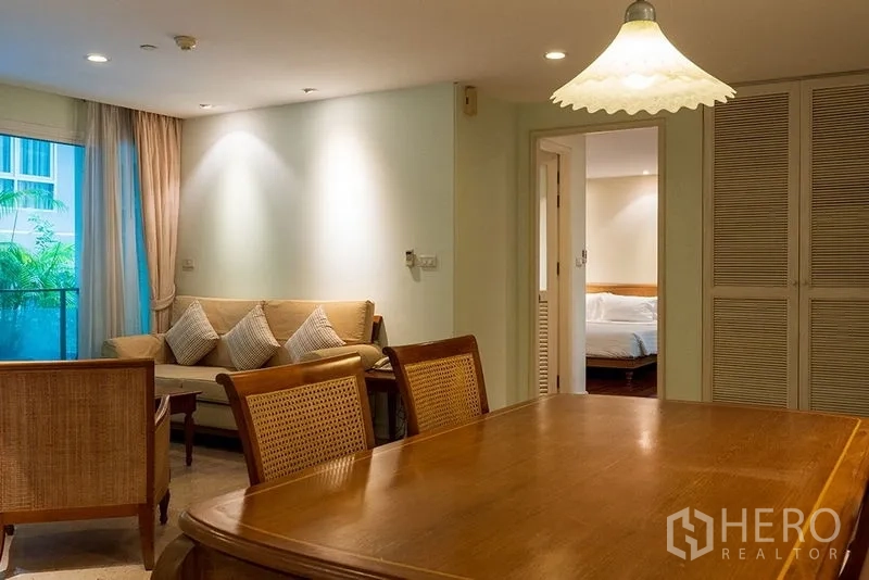 Apartment for rent in Bang Rak, Bangkok - Warm living and dining area with wooden table, rattan chairs, beige sofa and doorway to a bedroom.