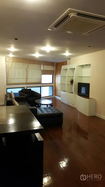 Condominium for rent in Khlong Toei, Bangkok - Lounge with black leather sectional sofa, built-in display shelves, large windows and cassette air conditioning.