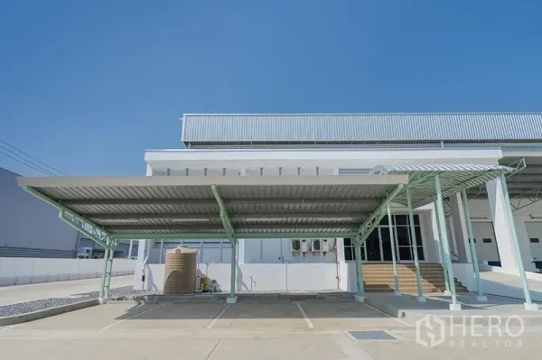 Warehouse for rent in Chon Buri - Warehouse exterior with covered parking, office entrance, and utility installations on a clear day.