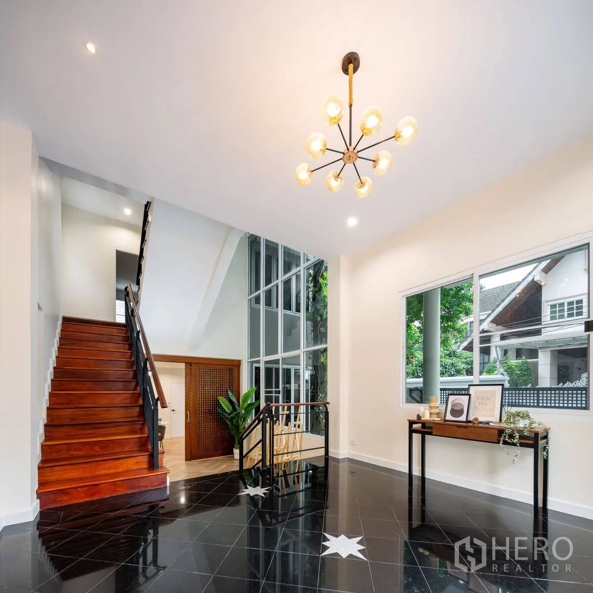 Detached House for rent or sale in Chatuchak, Bangkok - Double-height foyer with chandelier, glossy black flooring, large windows, and timber staircase.