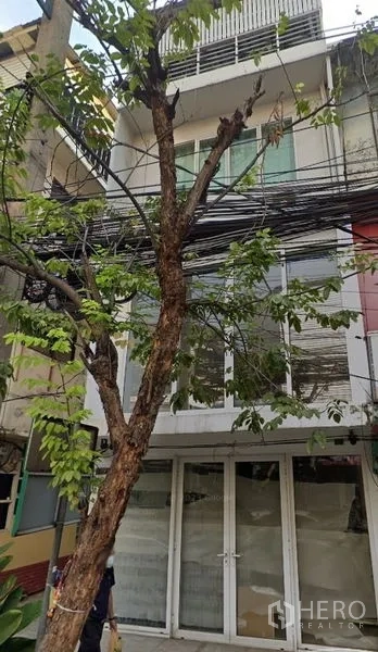 Shophouse for sale in Watthana, Bangkok - Three-storey shophouse facade with glass doors, a tree, and overhead utility cables in Watthana, Bangkok.
