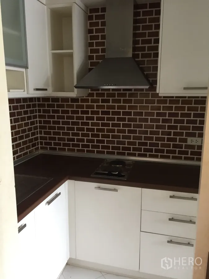Condominium for sale in Khlong Toei, Bangkok - Compact L-shaped kitchen with white cabinetry, brick backsplash, stainless range hood and cooktop.