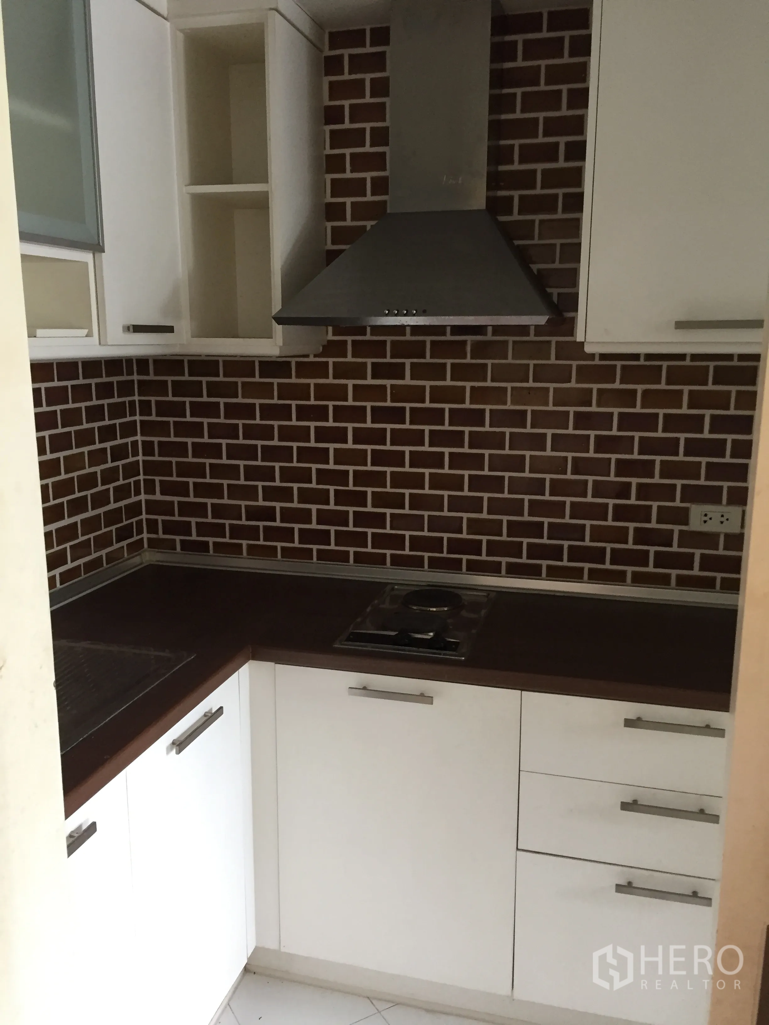 Condominium for sale in Khlong Toei, Bangkok - Compact L-shaped kitchen with white cabinetry, brick backsplash, stainless range hood and cooktop.