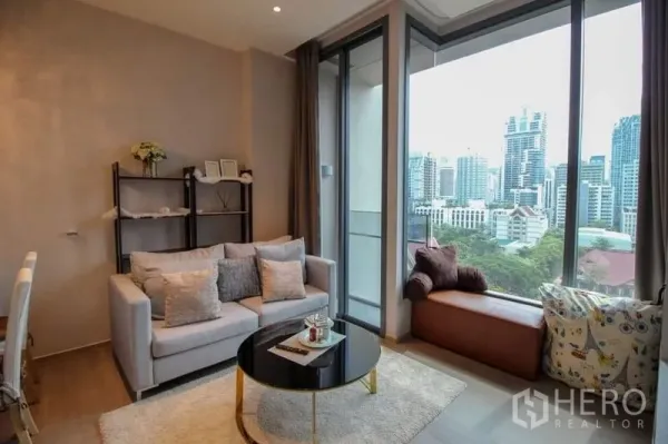 Condominium for sale in Watthana, Bangkok - Living room with window seat and floor-to-ceiling glass leading to the balcony, framing a city view.