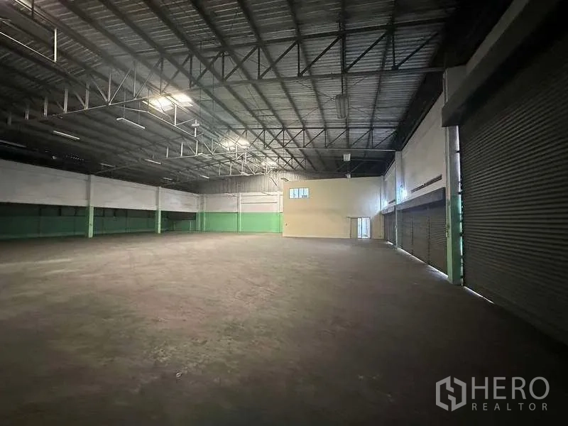 工厂 出租在Nonthaburi - 大型仓库大厅，钢桁架高挑屋顶、水泥地面与多道卷帘装卸门。