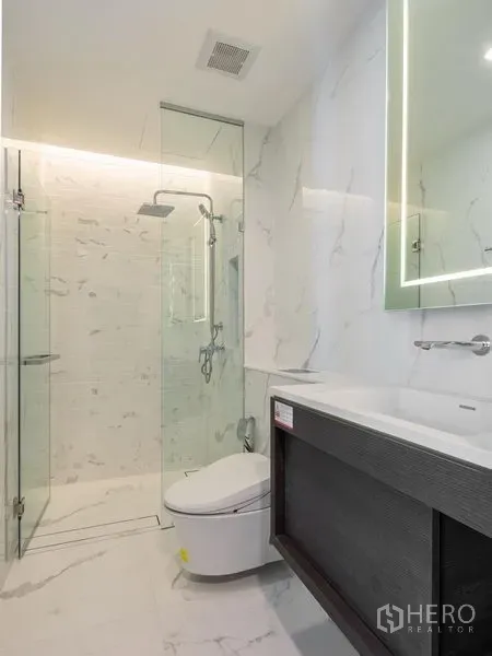 Condominium for rent in Khlong Toei, Bangkok - Bathroom with glass-enclosed rain shower, backlit mirror, and white marble-style tiles.