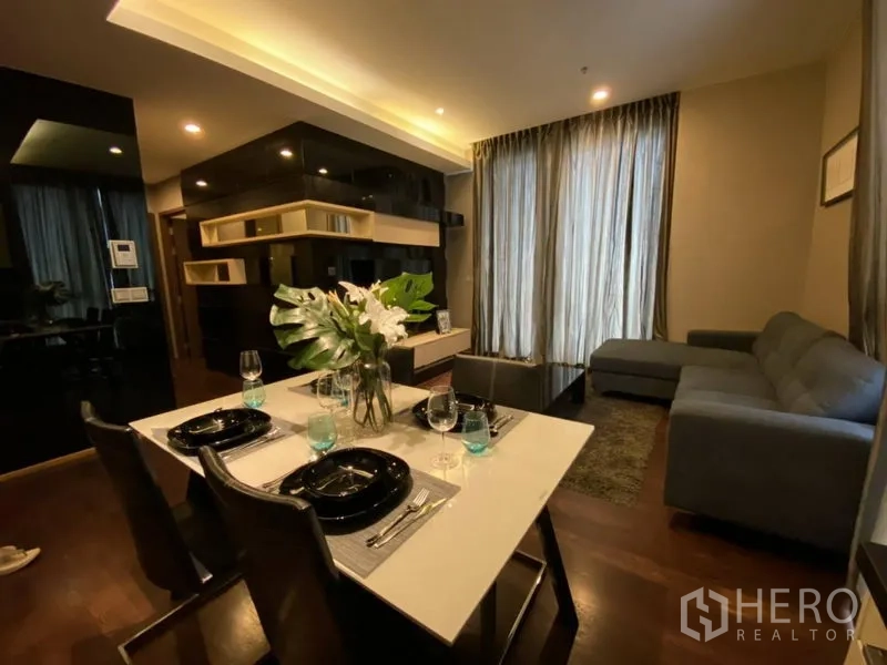 公寓 出租在Watthana, Bangkok - 通罗公寓的客餐厅一体空间，L 型沙发、内置置物架、木地板与落地窗帘。