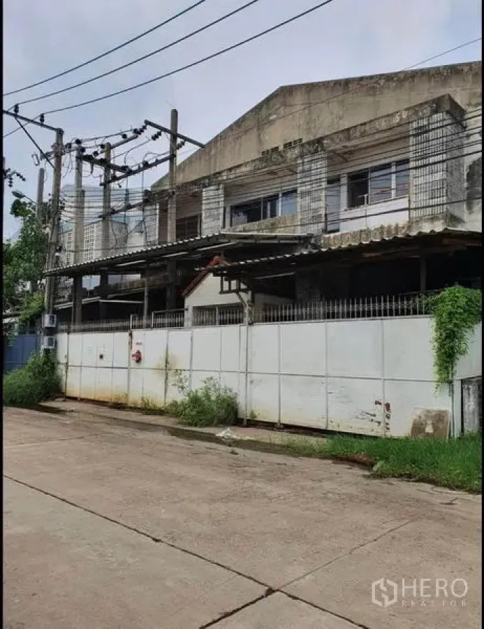 Warehouse for rent in Phra Samut Chedi, Samut Prakan - Street view of a two‑storey industrial warehouse behind a high concrete fence with gated entry and overhead power lines.
