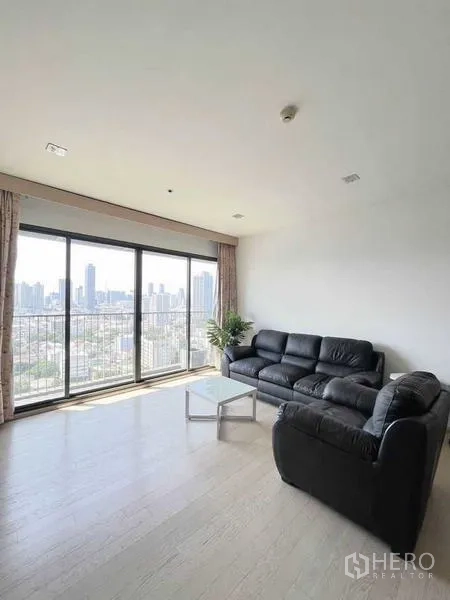 Condominium for sale - Bright Thonglor condo living room with black leather sofas, floor-to-ceiling windows, and panoramic Bangkok skyline view.