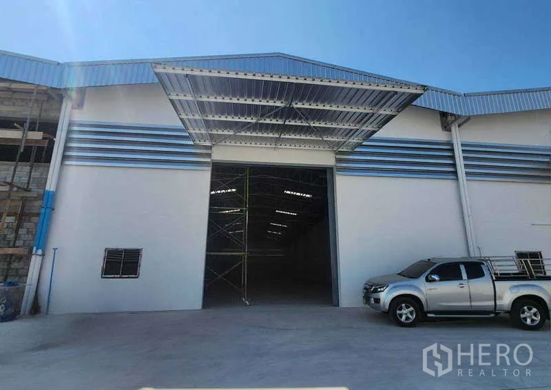 คลังสินค้า ให้เช่าในSam Phran, Nakhon Pathom - ด้านหน้าโกดังสร้างใหม่ มีหลังคาคุ้มพื้นที่โหลด ประตูทางเข้าขนาดใหญ่ และลานคอนกรีตพร้อมรถกระบะ