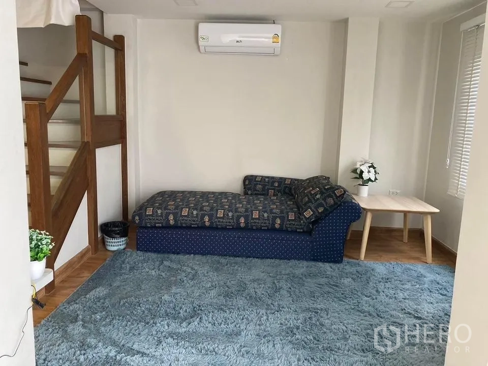 บ้าน ให้เช่าในKhlong Toei, Bangkok - มุมนั่งเล่นอบอุ่น โซฟาเดย์เบดสีน้ำเงินข้างบันไดไม้ พร้อมพรมผืนใหญ่