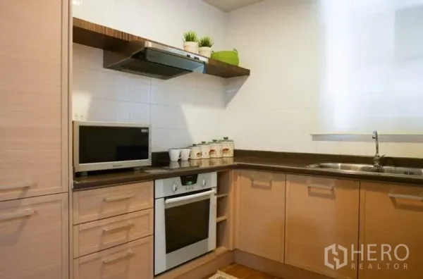 Condominium for rent in Pathum Wan, Bangkok - L-shaped kitchen with wood cabinetry, stone counters, oven, range hood, microwave and sink.