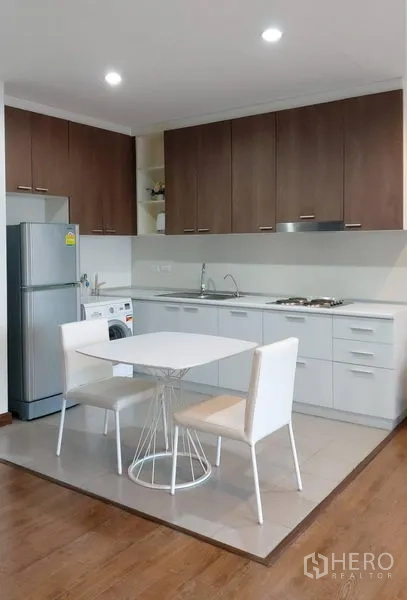 Condominium for rent in Phra Khanong, Bangkok - Compact kitchen with wood cabinets, hob, sink, refrigerator, washing machine and a dining table for two.