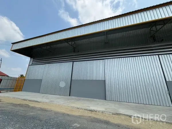 货栈 出租在Mueang Pathum Thani, Pathum Thani - 金属外墙仓库正面，宽幅滑动门与深型雨棚，前方装卸区域，蓝天映衬。