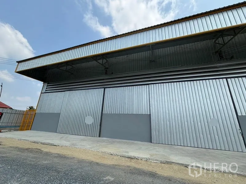 货栈 出租在Mueang Pathum Thani, Pathum Thani - 金属外墙仓库正面，宽幅滑动门与深型雨棚，前方装卸区域，蓝天映衬。
