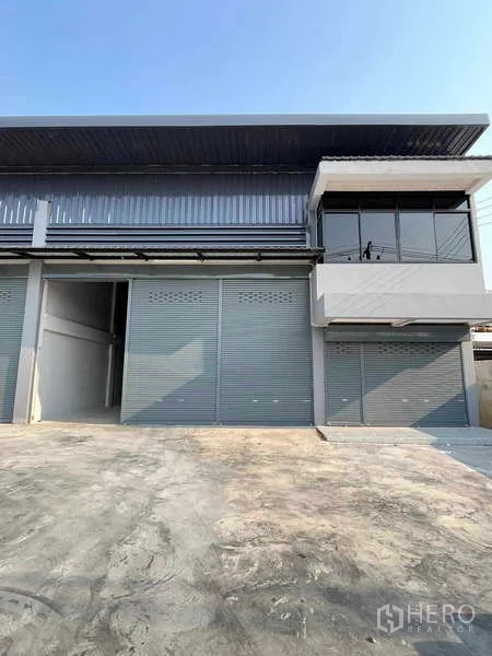 Warehouse for rent in Phra Samut Chedi, Samut Prakan - Front view of the warehouse showing a high metal roof, several roller shutter bays, and a side access opening for loading.