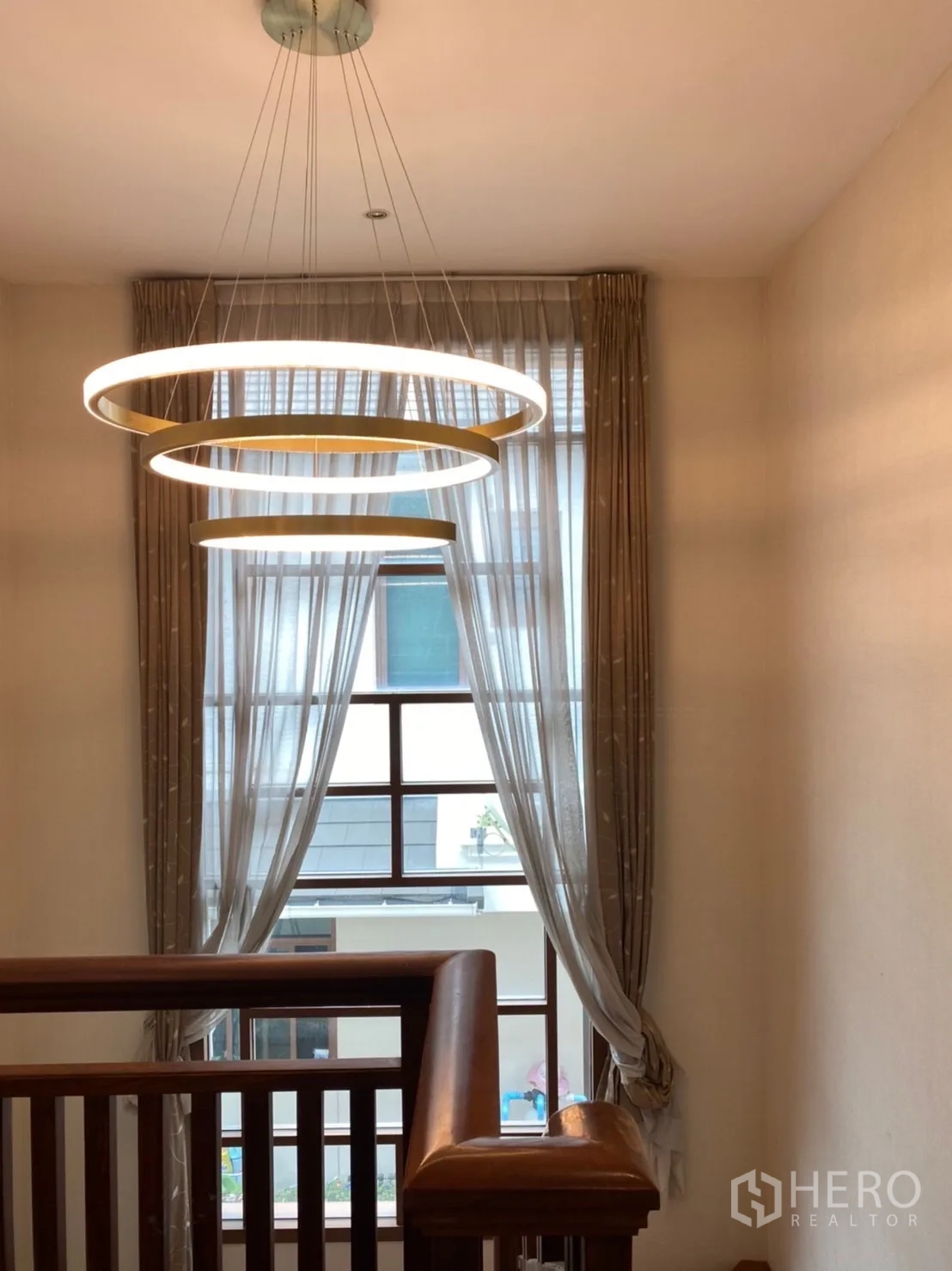Detached House for rent in Bang Phli, Samut Prakan - Stairwell with wooden handrail and three-ring chandelier beside a tall curtained window.