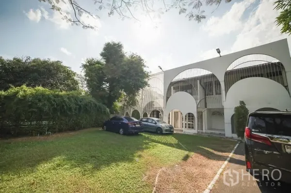 Detached House for rent in Huai Khwang, Bangkok - Front exterior with arched facade, large lawn parking area and mature trees.