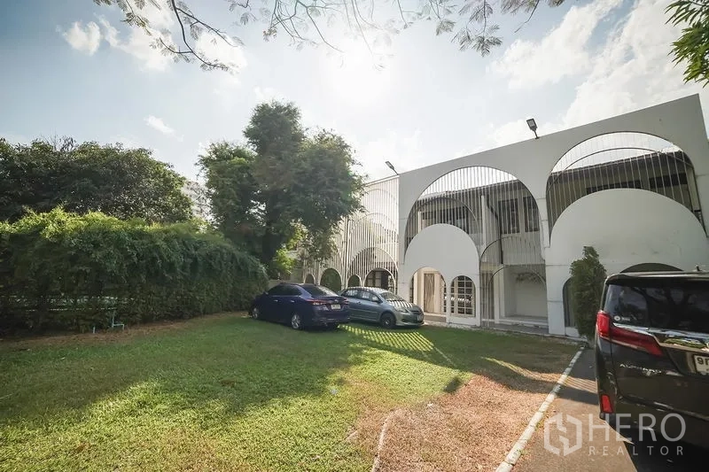 Detached House for rent in Huai Khwang, Bangkok - Front exterior with arched facade, large lawn parking area and mature trees.