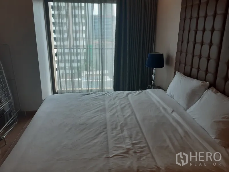 Condominium for rent in Bangkok - Bedroom with king bed, padded headboard and city view through tall window and curtains.
