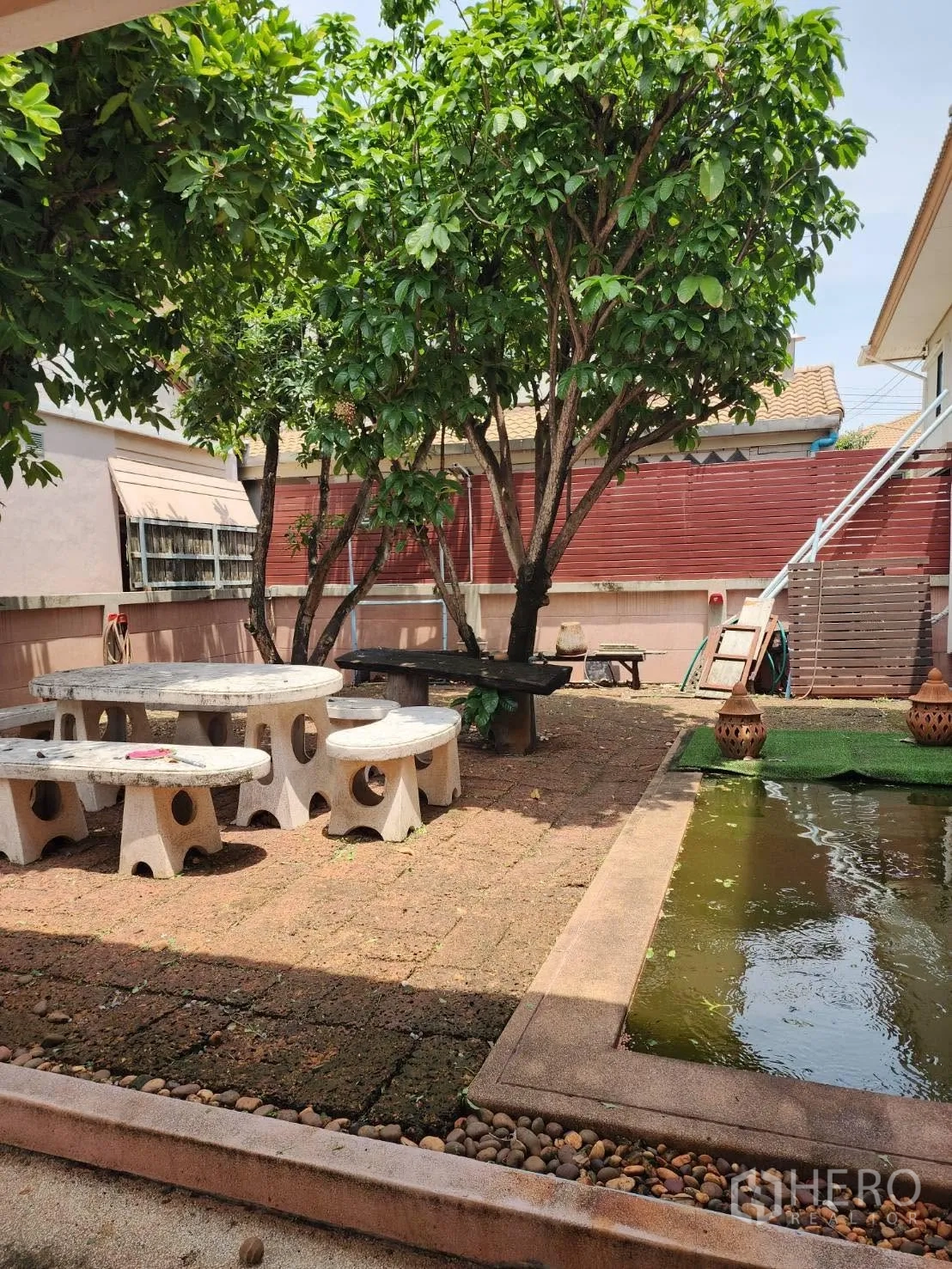 Detached House for rent in Lam Luk Ka, Pathum Thani - Backyard with stone picnic table set, paved patio, and a small fish pond among trees.