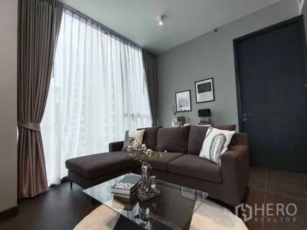 Condominium for rent in Bang Rak, Bangkok - Cozy lounge featuring an L-shaped sofa, tall draped windows, and framed wall art.
