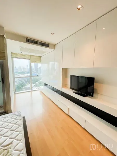 Condominium for rent in Khlong Luang, Pathum Thani - Bedroom with long built-in cabinet, TV, light wood floor, and large window offering a city view.