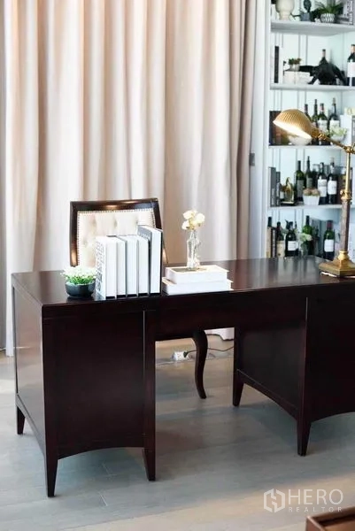 คอนโด ให้เช่า - มุมโต๊ะทำงานไม้สีเข้มพร้อมหนังสือ โคมไฟทองเหลือง และชั้นเก็บของในตัวหลังผ้าม่าน