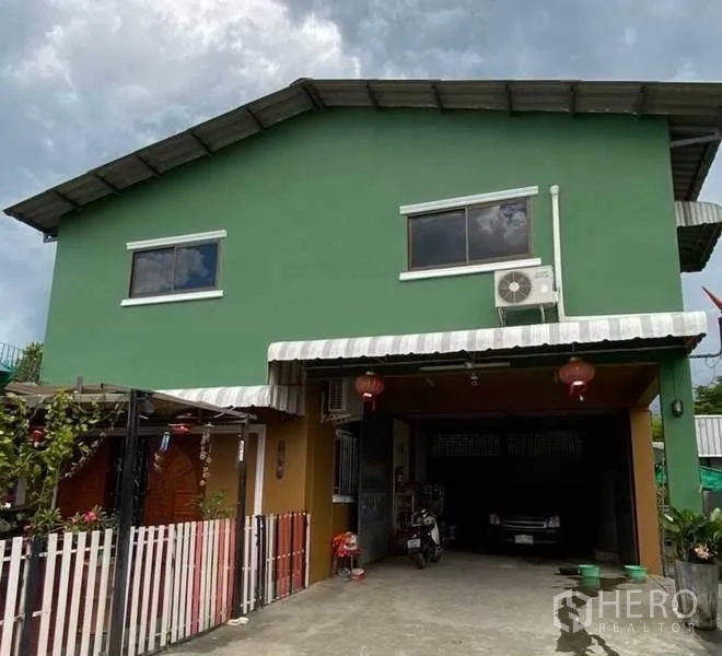 Warehouse for rent or sale in Pak Kret, Nonthaburi - Front view of a green two-storey building with striped awning, driveway parking and a wall-mounted AC unit.