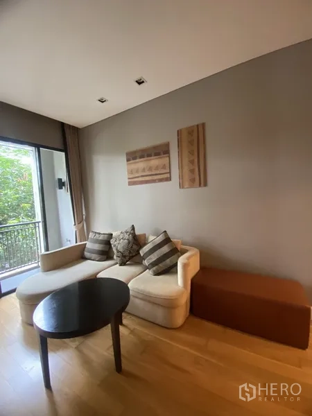 Condominium for rent in Watthana, Bangkok - Cozy living room with L-shaped sofa, coffee table, and sliding glass door to a leafy balcony.