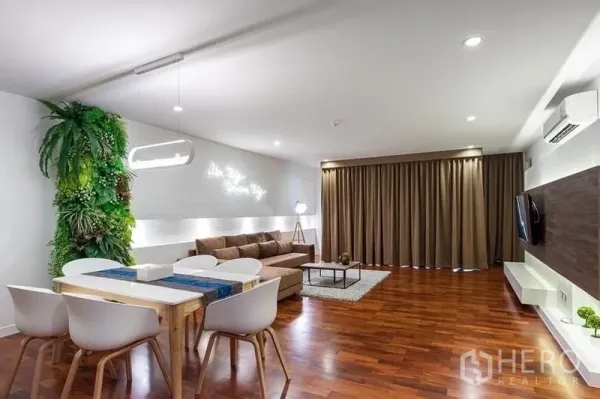 คอนโด ให้เช่าในPathum Wan, Bangkok - พื้นที่นั่งเล่นและรับประทานอาหารแบบโอเพ่นแปลน พื้นไม้ โซฟาสีน้ำตาล ผนังสวนแนวตั้ง และผ้าม่านสูงเต็มผนัง