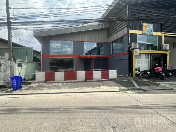 Warehouse for rent in Suan Luang, Bangkok - Wider view of the warehouse front and neighboring units with on-street parking and power lines.