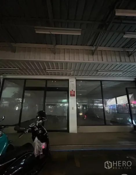 零售空间 出租在Suan Luang, Bangkok - 有顶人行道前的玻璃门店，夜间照明，停放有摩托车。