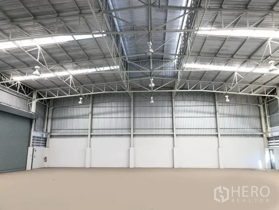 工厂 出租或出售在Bang Sao Thong, Samut Prakan - 高挑厂房仓库内景，钢结构屋架、天窗与高天棚灯，旁有卷帘门。