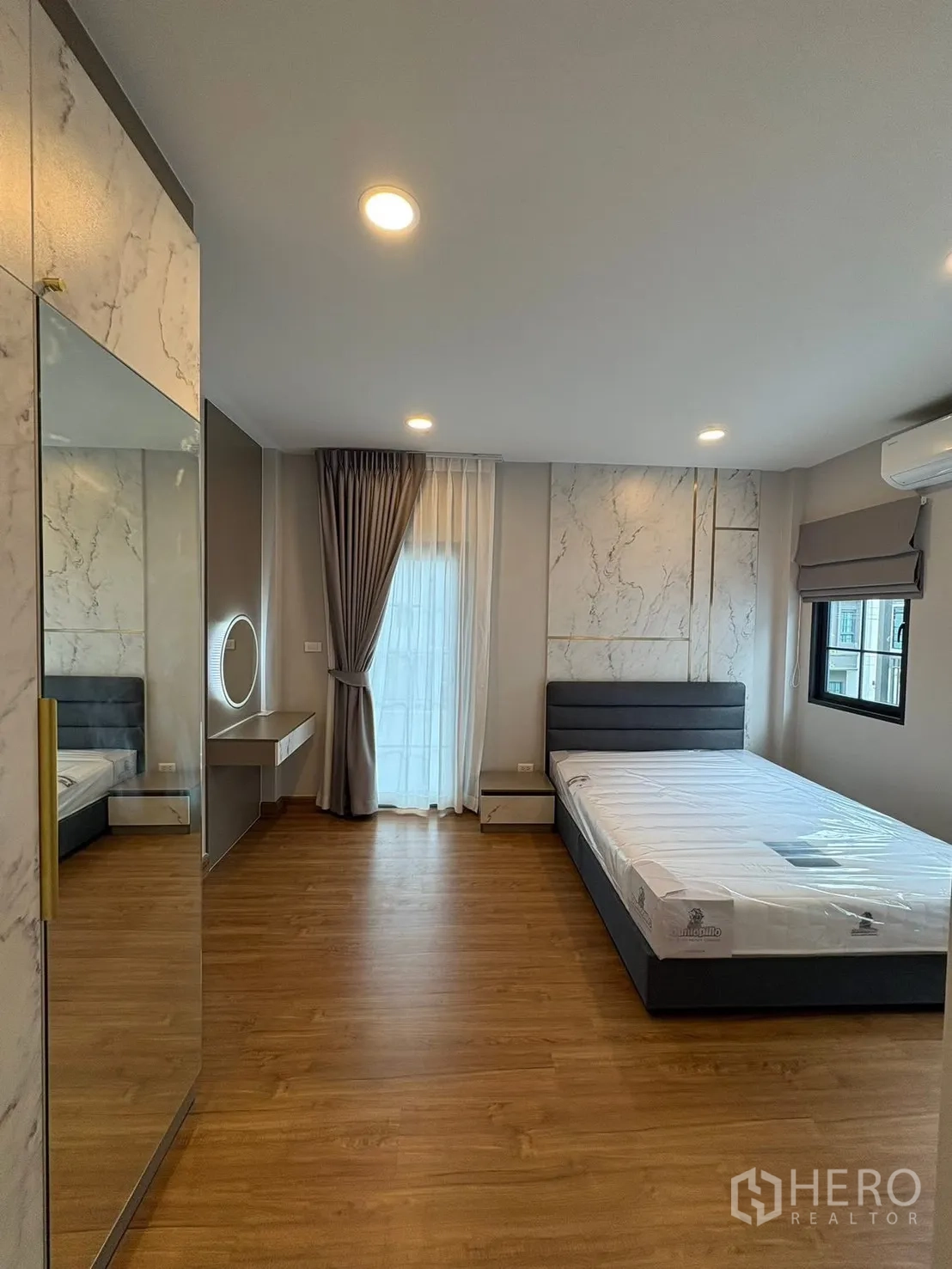 Detached House for rent in Bang Phli, Samut Prakan - Spacious bedroom with bed, marble-pattern wall panels, built-in mirror wardrobe, vanity shelf and wood flooring.