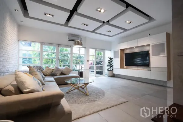 Townhouse for rent in Khlong Toei, Bangkok - Bright living room with L-shaped sofa, coffered ceiling, built-in TV wall and garden-view windows in a Bangkok townhouse.