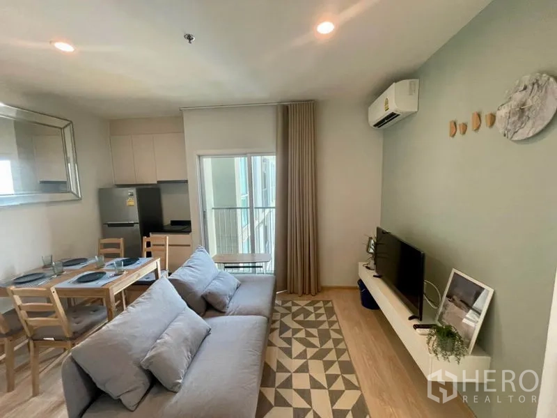 Condominium for rent in Huai Khwang, Bangkok - Bright living room with gray sofa, TV on floating shelf, balcony door and dining-kitchen.