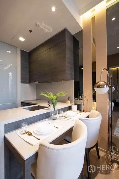 Condominium for rent in Watthana, Bangkok - Kitchen peninsula with two dining chairs, floor lamp and dark wood cabinets.