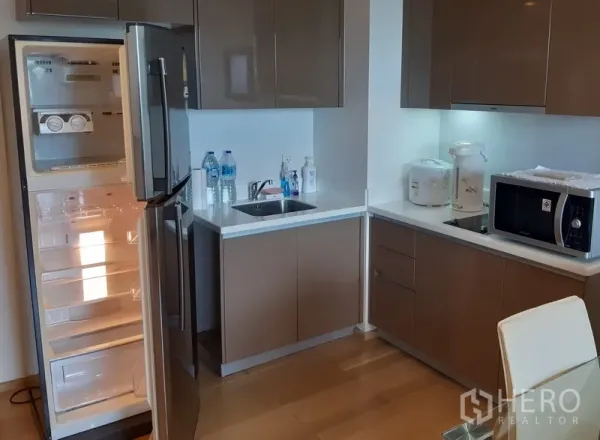 Condominium for rent in Khlong Toei, Bangkok - Modern L-shaped kitchen in a Phra Khanong condo with glossy cabinets, sink, microwave and an open stainless-steel fridge.