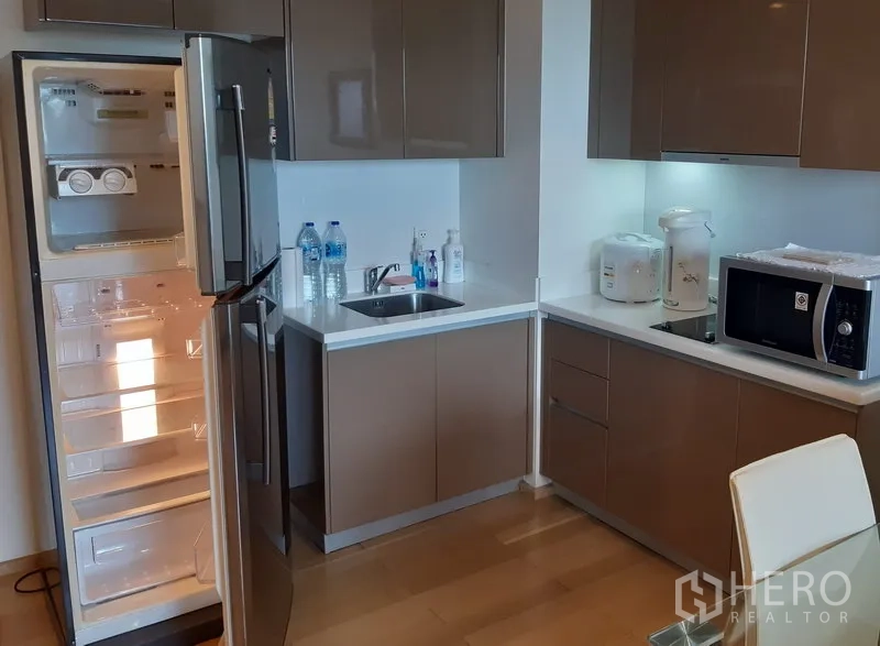 Condominium for rent in Khlong Toei, Bangkok - Modern L-shaped kitchen in a Phra Khanong condo with glossy cabinets, sink, microwave and an open stainless-steel fridge.