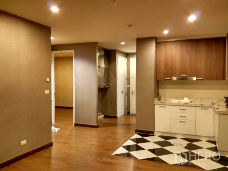 Condominium for rent in Phra Khanong, Bangkok - Open-plan living area with recessed lighting, warm wood floors, and a built-in kitchen with white cabinets and a black-and-white tiled cooking zone.