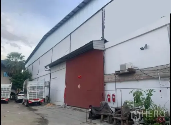 Warehouse for rent in Sai Noi, Nonthaburi - Large white warehouse exterior with high roof, red sliding door, fire extinguishers and truck loading area.