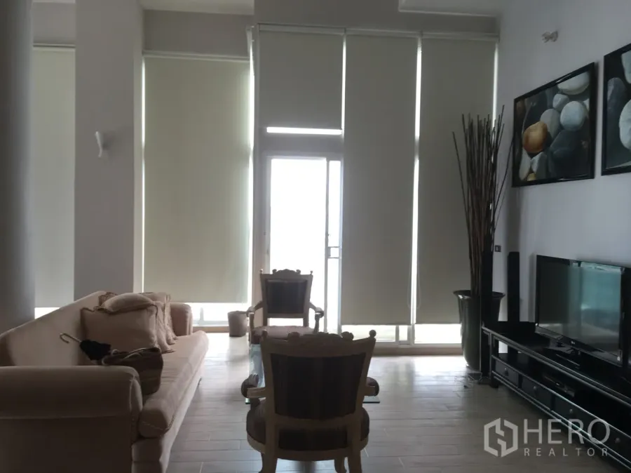 คอนโด ให้เช่าหรือขายในWatthana, Bangkok - ห้องนั่งเล่นเพดานสูง โปร่งสว่าง กระจกเต็มผนัง ม่านม้วน โซฟา และชั้นวางทีวี เปิดออกสู่ระเบียง