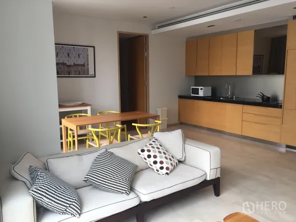 公寓 出租在Bang Rak, Bangkok - 客厅沙发与抱枕，旁边是极简木纹一字型厨房和餐桌椅。