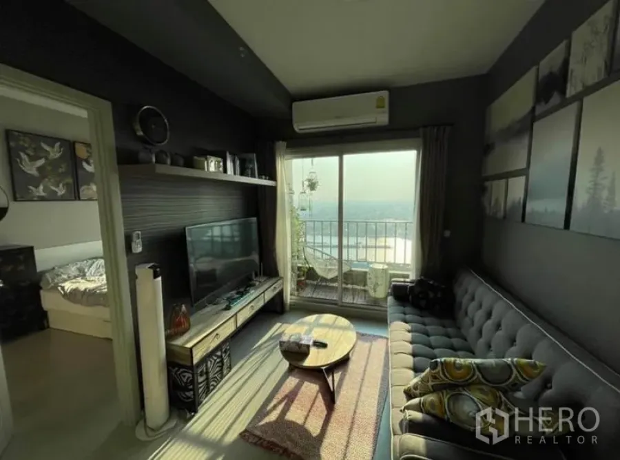 Condominium for sale in Bang Sue, Bangkok - Cozy living room with gray sofa, TV console and balcony opening to a wide river view.