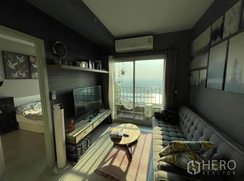 Condominium for sale in Bang Sue, Bangkok - Cozy living room with gray sofa, TV console and balcony opening to a wide river view.