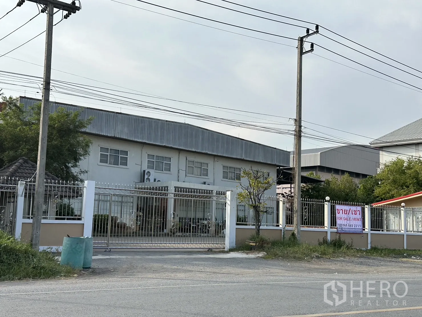 โรงงาน ให้เช่าหรือขายในLam Luk Ka, Pathum Thani - หน้าทางเข้าโรงงานพร้อมรั้วเหล็ก อาคารสำนักงานสองชั้น และสายไฟริมถนน ในลำลูกกา