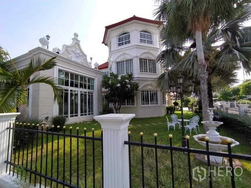 Detached House for sale in Phra Samut Chedi, Samut Prakan - Grand three‑storey detached house with red roof, large windows, palm‑lined lawn, garden seating and fountain behind a black fence.