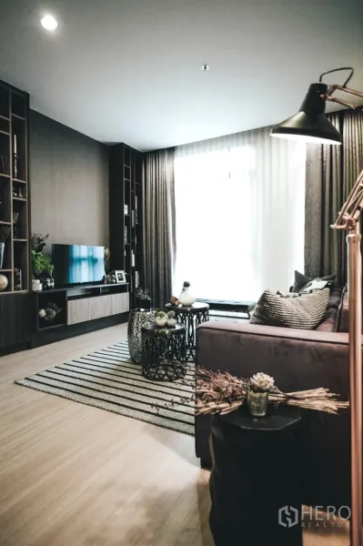 Condominium for rent in Huai Khwang, Bangkok - Cozy living room with large window, built-in shelving, sofa, striped rug, and nested coffee tables.