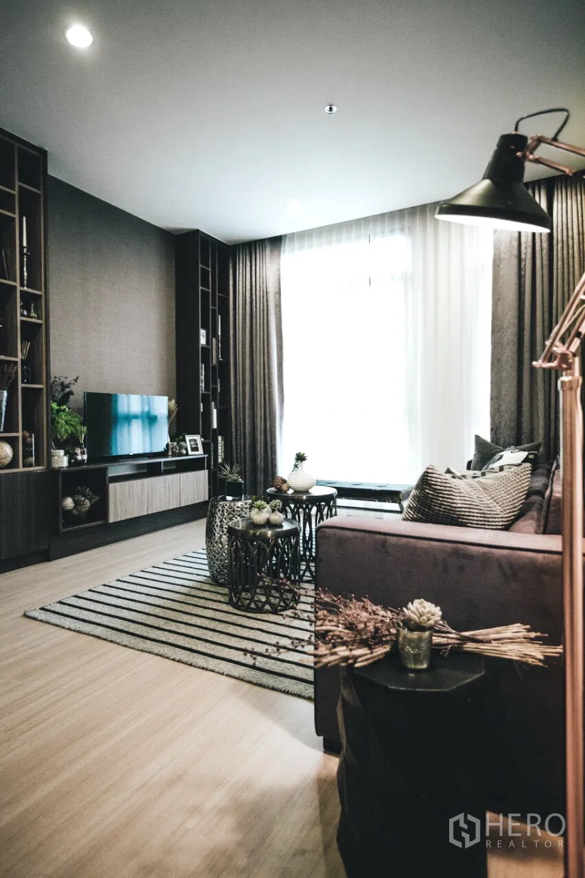 Condominium for rent in Huai Khwang, Bangkok - Cozy living room with large window, built-in shelving, sofa, striped rug, and nested coffee tables.